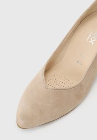 Beige mokkasiner med spiss tå, V-formet utskjæring og perforert såle for pusteevne. Flattenytt såle og glatt tekstur.