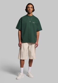 Grön oversized t-shirt med broderad logotyp, tillsammans med beige knälånga shorts. Vita sneakers och strumpor kompletterar outfiten.