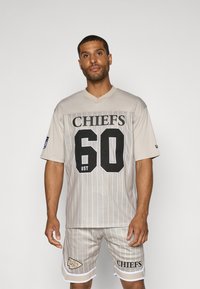 Μπεζ πλέγμα φανέλα ποδοσφαίρου με μαύρο κείμενο "CHIEFS" και αριθμό "60", σε συνδυασμό με ταιριαστές βερμούδες με λευκές λεπτομέρειες και λογότυπο.