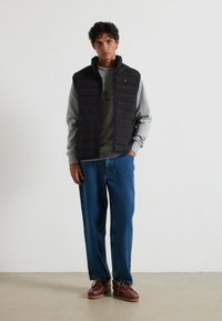 Schwarze gesteppte Weste über einem grauen Sweatshirt, kombiniert mit blauen Jeans und braunen Schuhen. Die Weste hat einen hohen Kragen und dezente Logodetails.