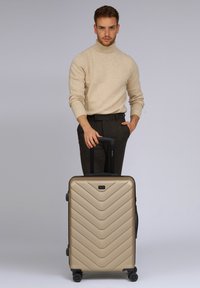 Hartschalenkoffer mit Chevron-Design in Metallic-Gold, schwarzen Rädern und einem ausziehbaren Griff, auf einem hellen Hintergrund platziert.