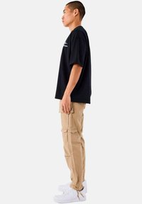 Sort oversized t-shirt, lyse brune cargo-bukser med lommer, hvide sneakers med strukturerede såler. Model står sidelæns mod en ensfarvet baggrund.