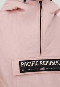 Lys pink hættepullover jakke med en sort patch med "PACIFIC REPUBLIC" og en lynlås. Lavet af et glat stof.