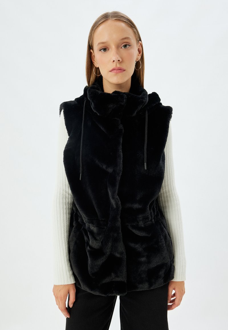 Schwarze Fleece-Weste mit hohem Kragen, Kordelzugverschluss und tailliertem Schnitt. Das Model trägt ein hellgraues, geripptes Langarmoberteil.