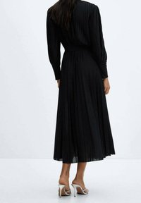 Robe midi plissée noire avec des manches longues et une taille ceinturée. Présente une texture lisse et une silhouette fluide, portée avec des escarpins argentés.