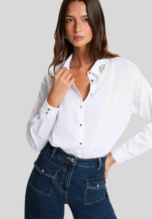 Mujer con camisa blanca de botones y detalle de pedrería en el cuello, y jeans de mezclilla oscura de tiro alto con grandes bolsillos frontales.