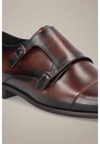 Brązowe skórzane buty na eleganckie wyjścia o gładkiej fakturze, wyposażone w dwie regulowane klamry, zaokrąglony nos oraz kontrastujący czarny pasek.