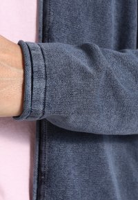 Dunkelblaues Langarmkleidungsstück aus strukturiertem Strickstoff, mit engem Bündchen und sichtbarer Nahtverzierung am Ärmel.