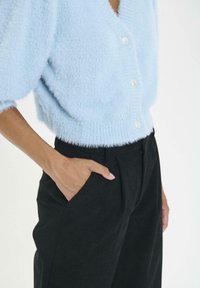 Persoon in een lichtblauwe fuzzy knooptrui en zwarte broek met één hand in de zak tegen een effen achtergrond.