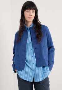 Blå jacka med krage och knappar, bärs över en ljusbl å randig skjorta. Outfiten kompletteras av mörka byxor.