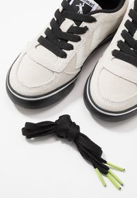 Beige Wildlederschuhe mit schwarzen Schnürsenkeln und schwarzer Gummisohle, ausgestattet mit einem weißen Logo und Akzenten, begleitet von schwarzen Schnürsenkeln mit grünen Spitzen.