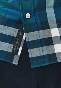 Blau-grün kariertes Kurzarmhemd mit weißen Akzenten, Knopfverschluss und einem schwarzen Etikett mit der Aufschrift "Durable Goods Lot 082" am Saum.