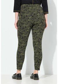 Persona vestita con leggings maculati neri e verde oliva e mocassini neri, in piedi contro un muro bianco, voltata di spalle.