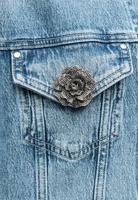 Τσέπη τζιν μπουφάν με μια υφασμάτινη, μεταλλική καρφίτσα σε σχήμα τριαντάφυλλου. Υφασμάτινο ύφασμα γαλάζιου χρώματος με ασημένιες λεπτομέρειες και χαρακτηριστικά σχέδια ραφής.