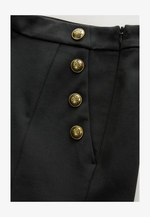 Musta hame, jossa on rakenteellinen vyötärö, neljä koristeellista kultanappia monimutkaisilla kuvioilla ja sivuvetoketju. Sileä, mattapintainen rakenne.