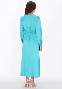 Langærmet, turkis satin kjole med en tætsiddende talje og flydende skørt. Har en rund halsudskæring og glat tekstur. Elegant design.