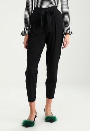 Femme portant un pantalon noir taille haute avec une ceinture nouée, une chemise rayée à manches longues et des talons noirs avec des détails en plumes vertes.
