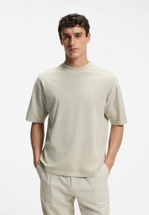 Jeune homme portant un T-shirt beige clair à manches courtes et un pantalon assorti, debout les mains dans les poches sur fond blanc.