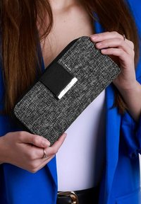 Peněženka z texturované látky v černé a šedé barvě se zipovým uzávěrem, která má černou elastickou pásku a stříbrnou logo destičku na přední straně.
