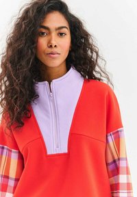 Czerwony sweter z lawendowym kołnierzem i zamkiem; wzorzyste, kratkowane rękawy w kolorach pomarańczowym, różowym, fioletowym i kremowym; miękki materiał, luźny krój.