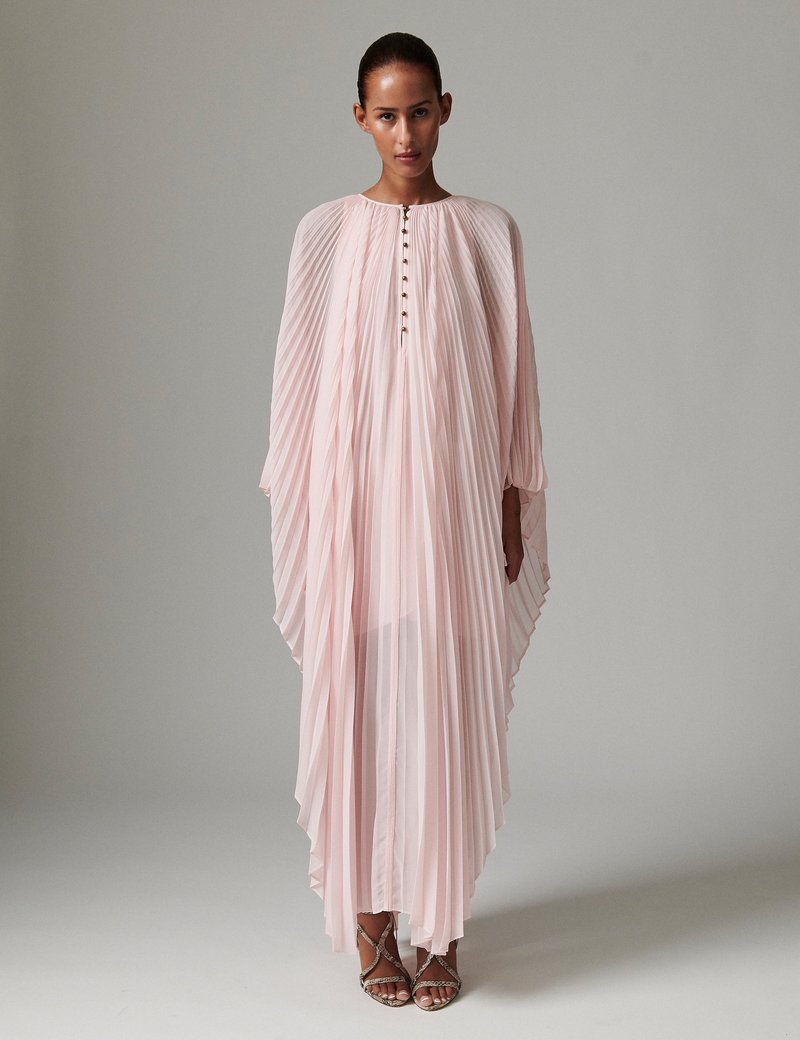 Robe longue plissée en tissu rose doux avec un col haut et un devant boutonné. Caractérisée par des manches fluides semblables à une cape et une texture légère.