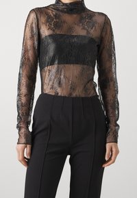 Top negro de encaje de mangas largas con una superposición transparente y una banda negra sólida a través del pecho. Presenta delicados patrones florales y un cuello alto.