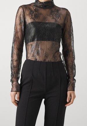 Černá krajková halenka s dlouhými rukávy a průhlednou vrstvou, s pevným černým páskem přes hruď. Obsahuje jemné květinové vzory a vysoký výstřih.