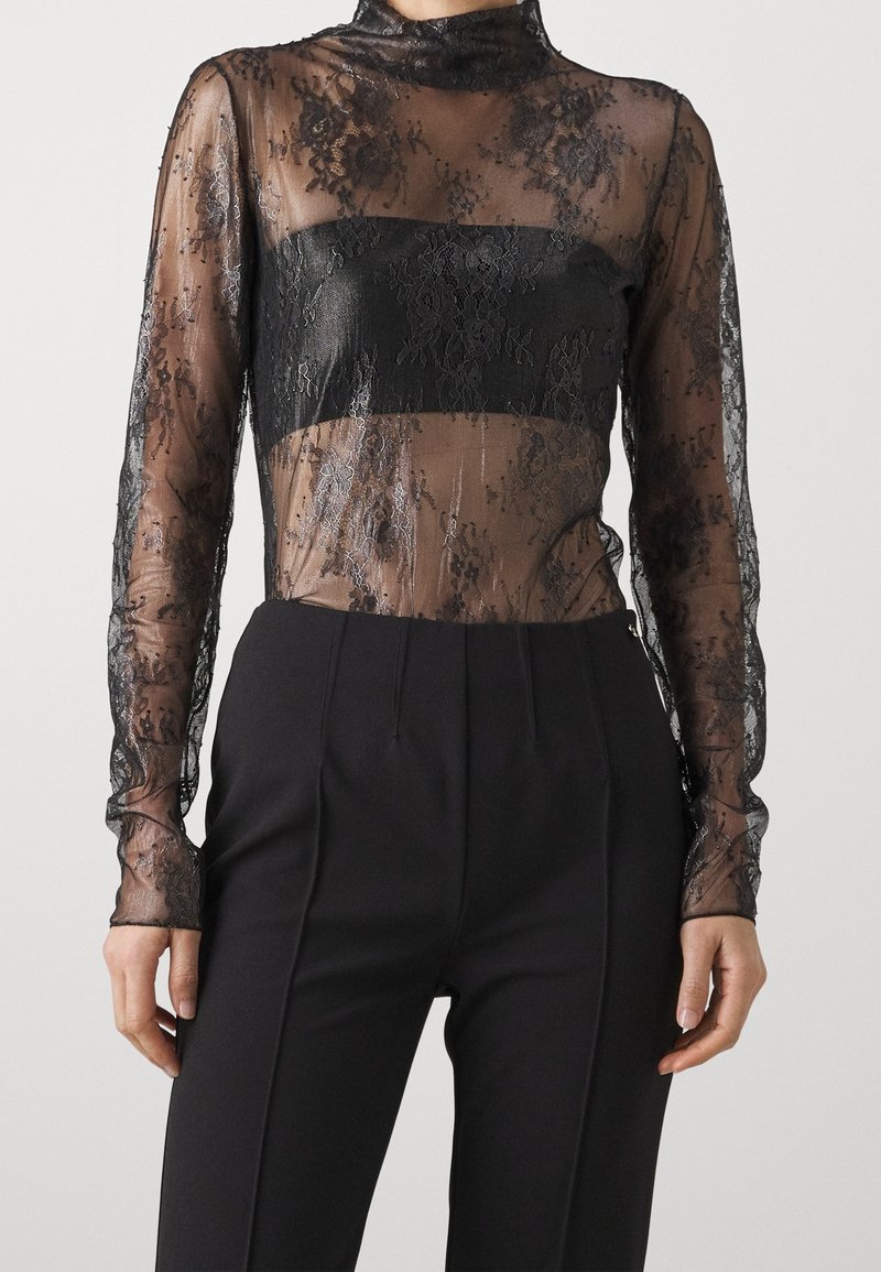 Top negro de encaje de mangas largas con una superposición transparente y una banda negra sólida a través del pecho. Presenta delicados patrones florales y un cuello alto.