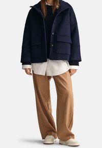 Giacca in misto lana navy con cappuccio, grandi tasche frontali e chiusura con bottoni, abbinata a una camicia bianca e pantaloni beige a gamba larga.