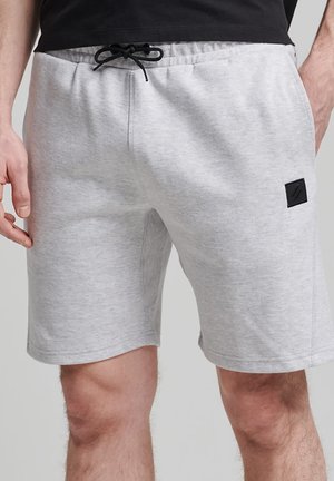 Mand iført lysegrå shorts med snøre og sort lap på venstre lår, kombineret med en sort skjorte, står med hænderne ved siden.