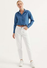 Camicia blu corta con bottoni, maniche lunghe e taschino sul petto, abbinata a pantaloni bianchi e sneakers bianche.