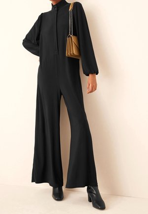Femme portant une combinaison noire à manches longues avec jambes larges, des bottines noires et un petit sac à main marron porté à l'épaule.