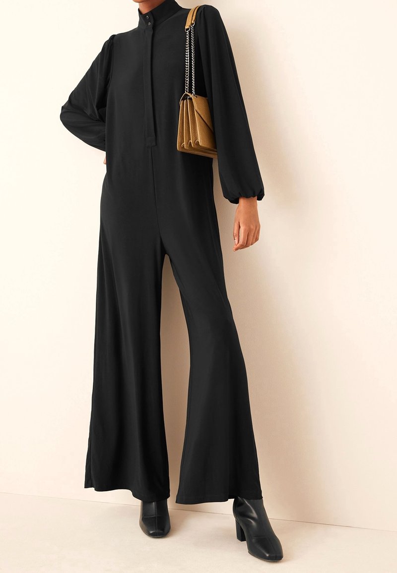Femme portant une combinaison noire à manches longues avec jambes larges, des bottines noires et un petit sac à main marron porté à l'épaule.