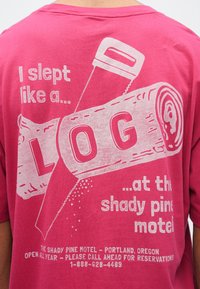Rosa bomullst-skjorte med grafisk design av en tømmerstokk, sag og tekst. Trykket er hvitt; skjorten har rund hals og korte ermer.
