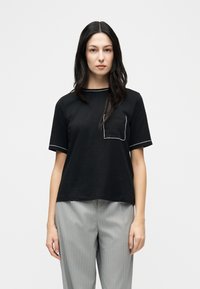 Γυναίκα με μακριά σκουρόχρωμα μαλλιά που φοράει μαύρο κοντομάνικο τοπ με λευκή επένδυση και τσέπη στο στήθος, καθώς και γκρι ριγωτό παντελόνι, στέκεται μπροστά σε λευκό φόντο.