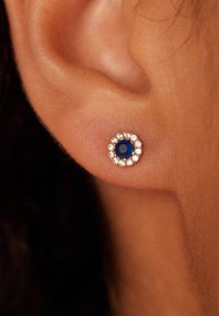 Ronde studoorbel met een centrale blauwe edelsteen omringd door kleine heldere kristallen, gezet in een zilverkleurige metalen basis. Gladde, gepolijste afwerking.