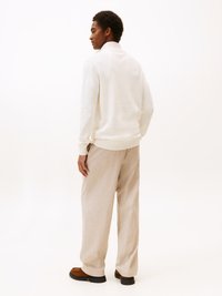 Felpa bianca con colletto alto, abbinata a pantaloni a gamba larga beige chiaro. Calzature marroni con suola nera. Design minimalista, colori neutri.