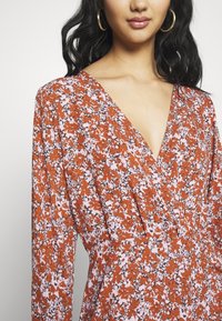 Femme portant une robe portefeuille à manches longues, à fleurs rouille et blanches, avec un décolleté en V et des boucles d'oreilles dorées en forme d'anneau, sur un fond uni.