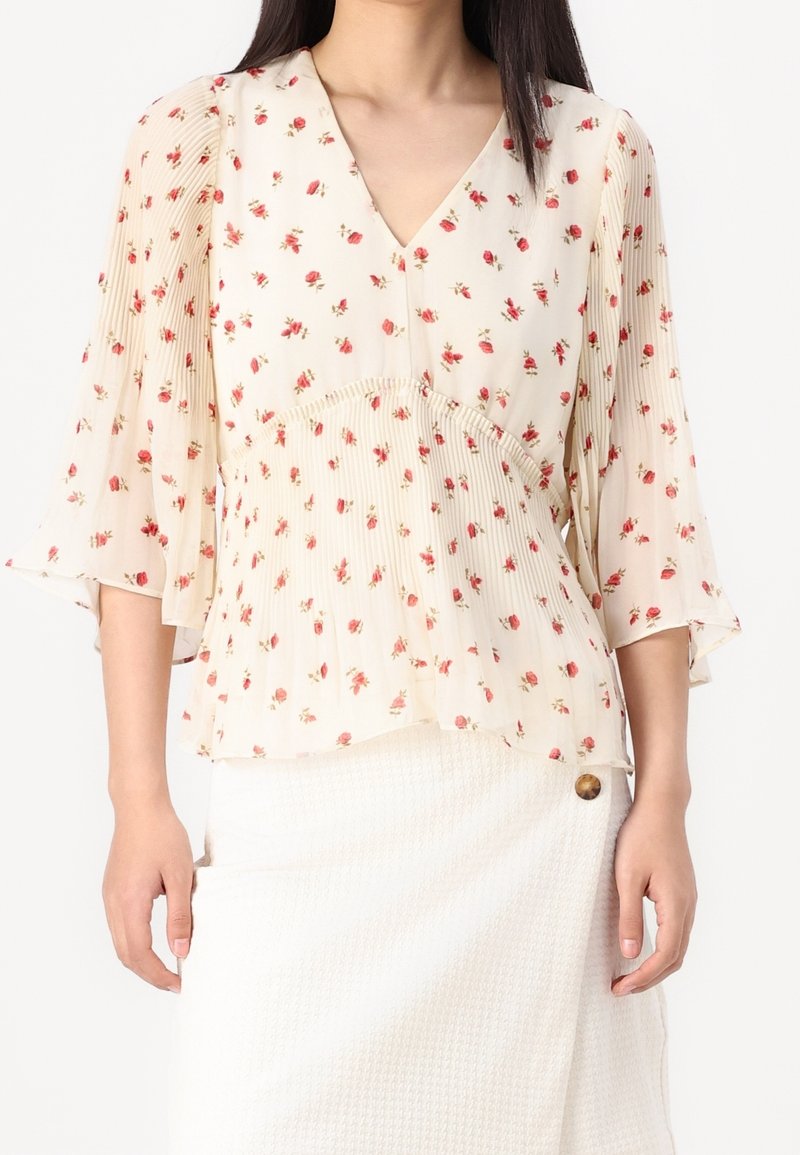 Blusa floral con un fondo crema claro, adornada con pequeñas flores rojas, escote en forma de V, textura plisada y mangas anchas y fluidas.
