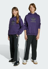 Un sweat à capuche violet avec un logo adidas noir, associé à un pantalon noir avec des bandes blanches sur les côtés et des baskets. Enfants en tenue décontractée.