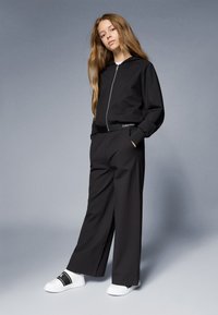 Fekete cipzáras kapucnis pulóver széles szárú fekete nadrággal párosítva, melynek derékpántján Calvin Klein logó található. A szettet fehér slip-on cipők egészítik ki.