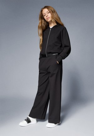 Crna zip-up dukserica uparena s crnim širokim hlačama, s Calvin Klein logotipom na pojasu. Bijele slip-on cipele upotpunjuju odjevnu kombinaciju.