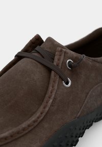 Scarpa in suede marrone con forma arrotondata, caratterizzata da lacci elasticizzati grigi, occhielli in argento e suola in gomma nera testurizzata.