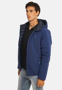 Homme portant une veste à capuche bleue sur un polo noir et un jean bleu clair, debout et regardant sur le côté sur un fond blanc.