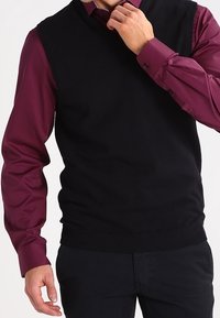 Veste en maille noire avec un col en V, portée sur une chemise bordeaux ajustée. La veste présente un ourlet côtelé et une texture lisse.