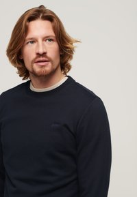 Felpa blu navy con colletto rotondo, caratterizzata da un colletto interno grigio chiaro e un piccolo logo ricamato sul petto. Materiale morbido e vestibilità regolare.