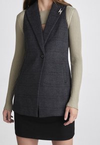 Gilet grigio scuro con motivo a righe verticali, chiusura a un bottone, rever a punta, indossato sopra un top oliva a coste, abbinato a una gonna nera.