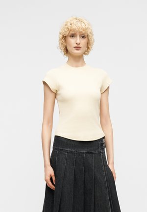 Γυναίκα με κοντά σγουρά ξανθά μαλλιά, που φοράει μπλούζα μπεζ με κοντά μανίκια και ψηλόμεση μαύρη πλισέ φούστα, στέκεται μπροστά σε απλό φόντο.