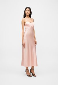 Naine, kes kannab heleroosa satiinist midi-kleit õhukeste rihmadega ja mustade kõrge kontsaga sandaalidega, seisab ühevärvilise tausta ees.