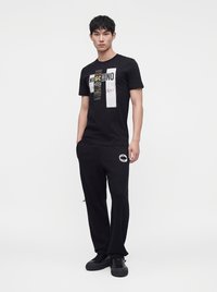 Νέος άνδρας που φοράει μαύρο γραφικό t-shirt Moschino, μαύρο παντελόνι φόρμας Moschino και μαύρα αθλητικά παπούτσια, στέκεται με τα χέρια στις τσέπες.